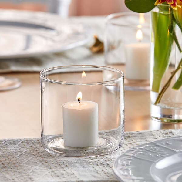 An Acopa cylindrical glass candle holder with a lit candle on a table.