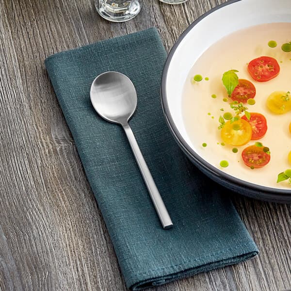 An Acopa Phoenix stainless steel bouillon spoon in a bowl of soup with tomatoes and basil on a table with a cloth.