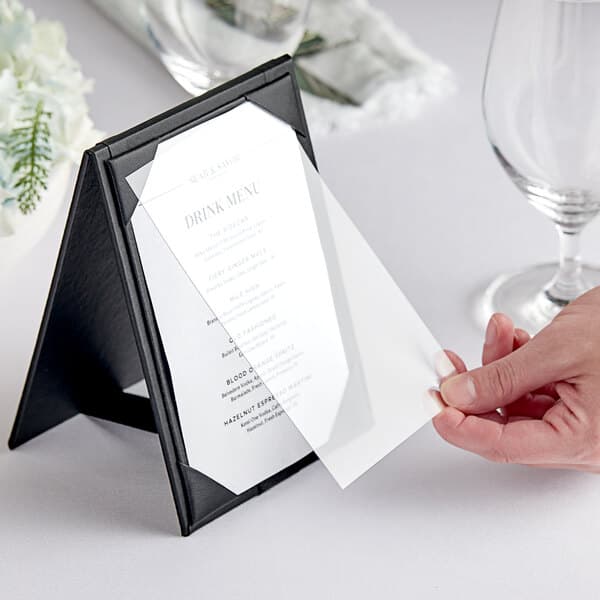 A close-up of a hand holding a menu card with a wine glass on a table.