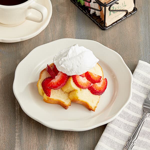 An Acopa ivory scalloped stoneware plate with a piece of cake topped with strawberries and whipped cream.