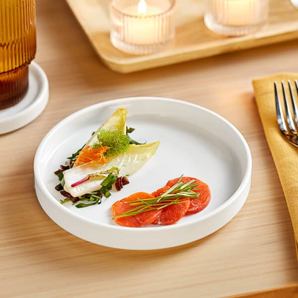 A plate of food on a wood table with a fork and knife on an Acopa Lore raised rim porcelain plate.
