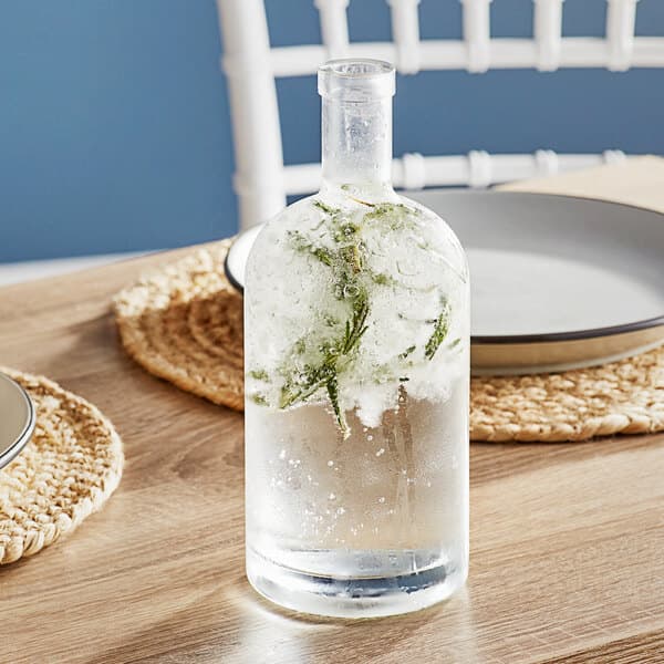 An Acopa clear glass bottle with ice and a sprig of mint on a table.