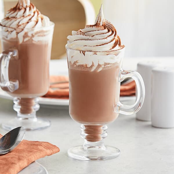 Two Acopa Select glass cups of hot chocolate with whipped cream on a table.
