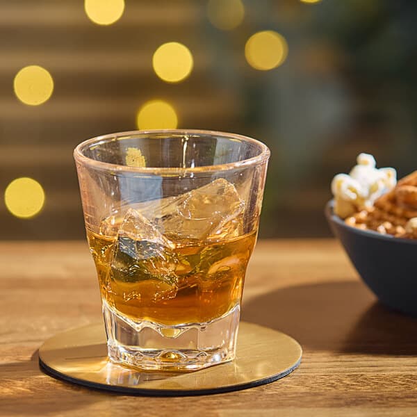 An Acopa Memphis rocks glass of amber liquid with ice on a coaster with a bowl of popcorn.