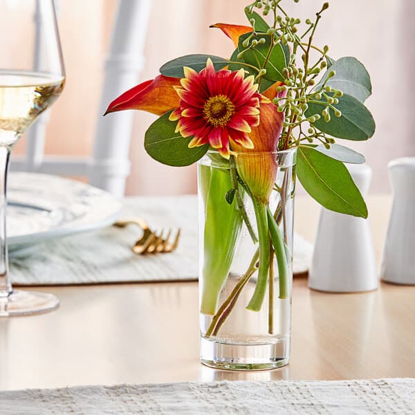An Acopa cylindrical glass bud vase with flowers and leaves on a table.
