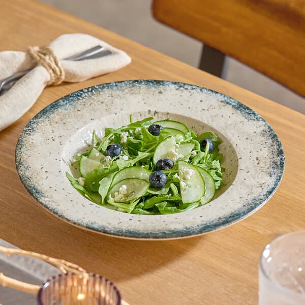 A round gray porcelain wide rim bowl filled with a green salad, cucumber slices, blueberries, and cheese crumbles.
