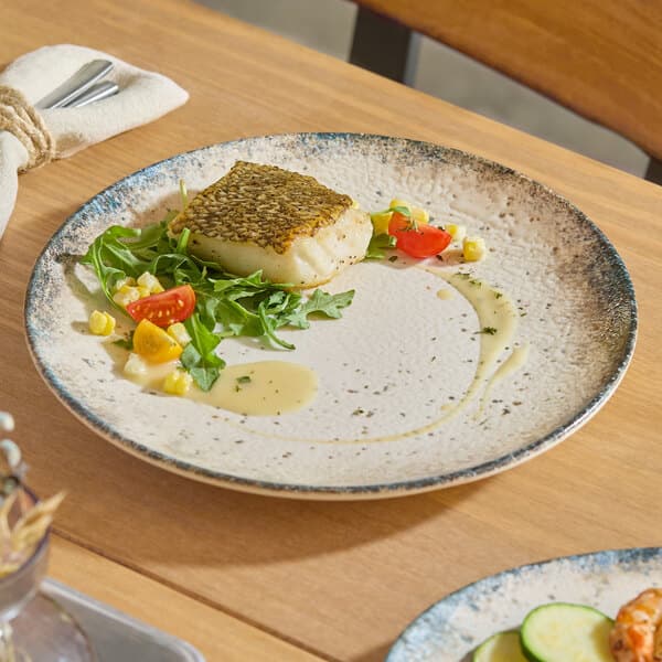 A round gray porcelain coupe plate with a speckled design, featuring a serving of fish, greens, and vegetables.