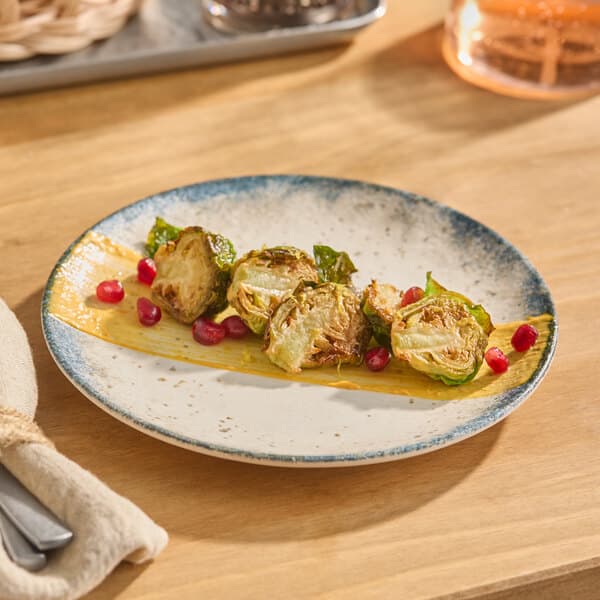A round gray porcelain coupe plate with a blue rim, featuring roasted Brussels sprouts, a yellow sauce, and pomegranate seeds.