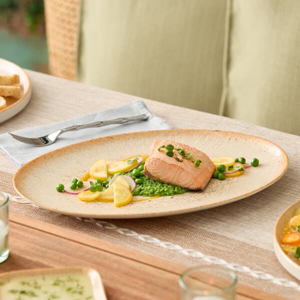 An oval beige porcelain platter displaying a serving of salmon with vegetables.