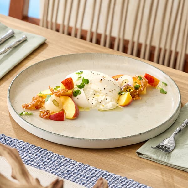 A round gray porcelain plate featuring a serving of burrata cheese with sliced peaches, prosciutto, and microgreens.