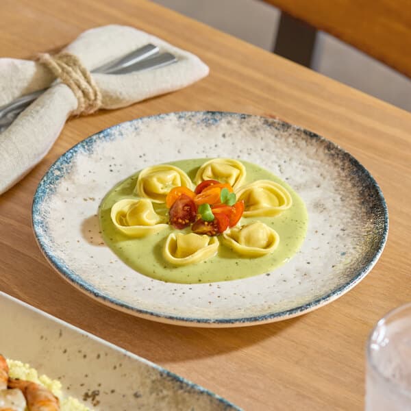 A round gray porcelain coupe plate with a speckled rim, featuring a serving of tortellini in a green sauce and garnished with cherry tomatoes.